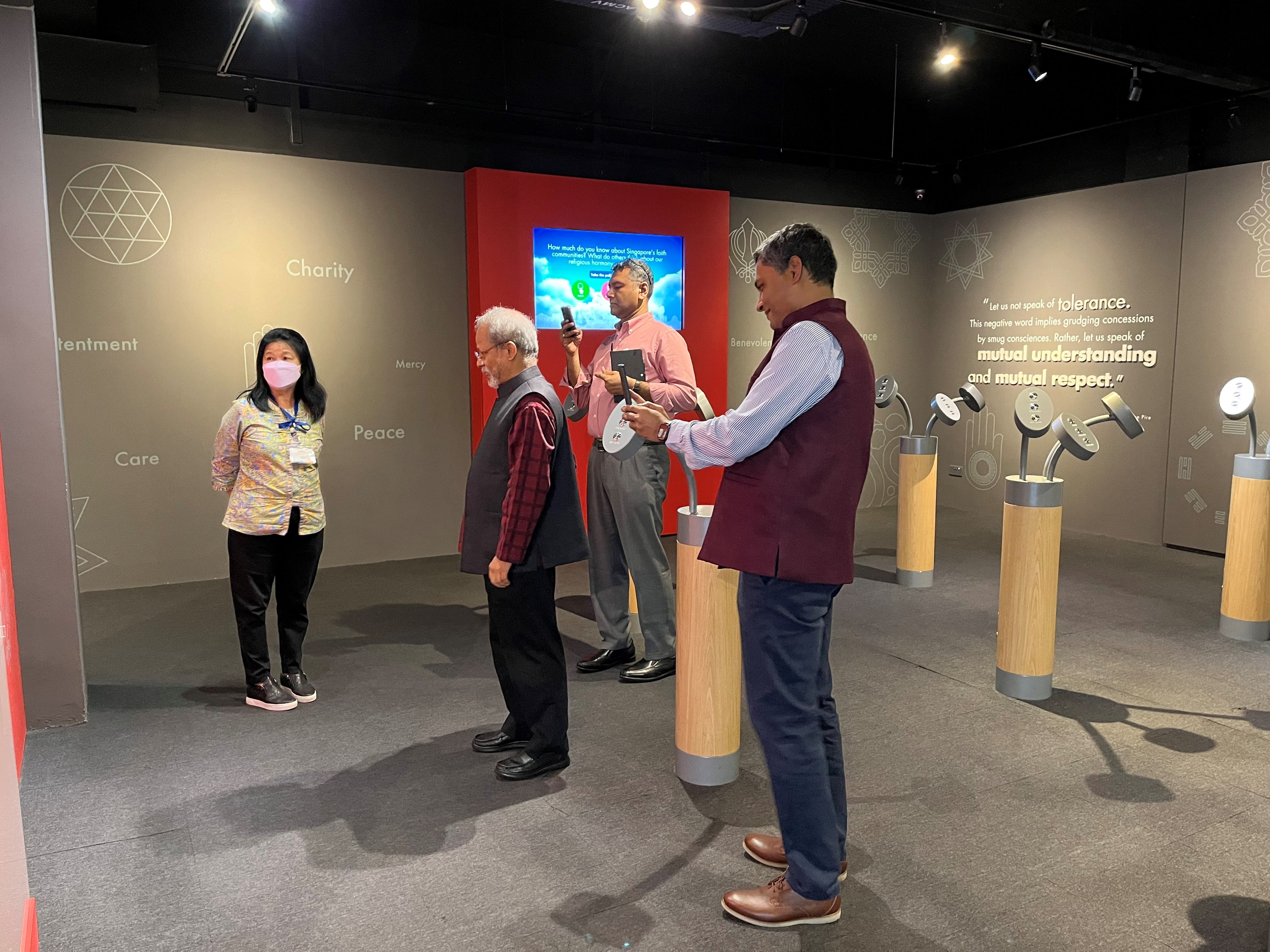 Four people stand in a museum exhibit with words like Charity, Peace, and Care on gray walls, with interactive displays.